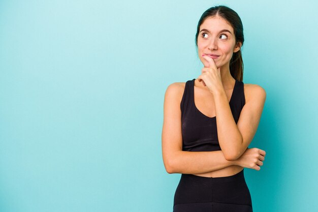 Young sporty caucasian woman isolated on blue background relaxed thinking about something looking at a copy space.