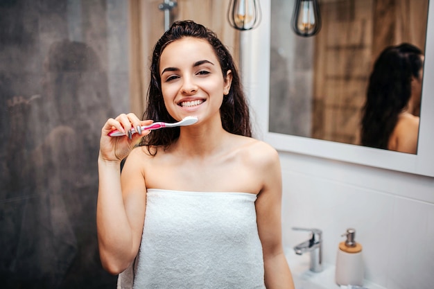 Young sporty dark-haired beautiful woman doing morning evening routine at mirror. She look on camera and smile. Model hold tooth paste with tooth brush close to mouth. Posing on camera.