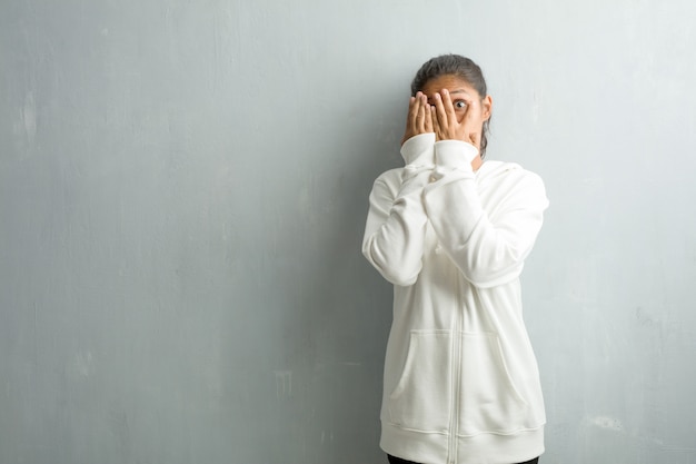 Young sporty indian woman against a gym wall feels worried and scared, looking and coverin