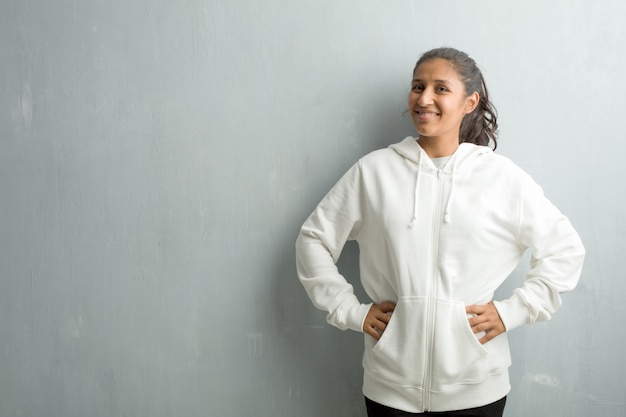 Young sporty indian woman against a gym wall with hands on hips, standing, relaxed and smi