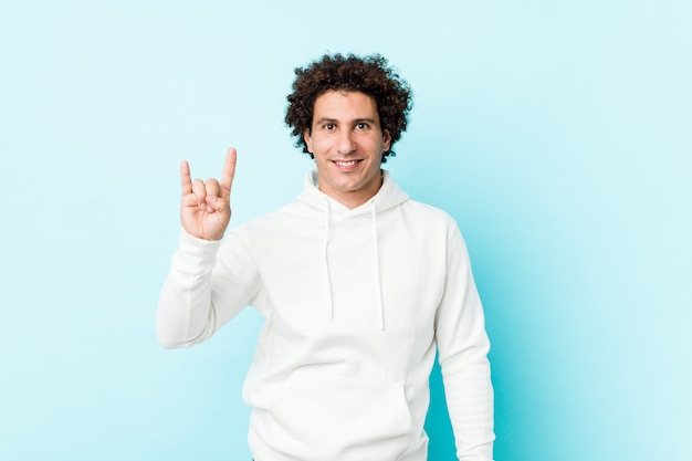 Young sporty man against a blue wall showing a horns gesture as a revolution concept.