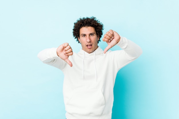 Young sporty man against a blue wall showing thumb down and expressing dislike.