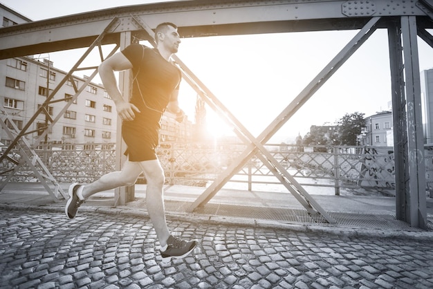 young sporty man jogging across the bridge at sunny morning in the city