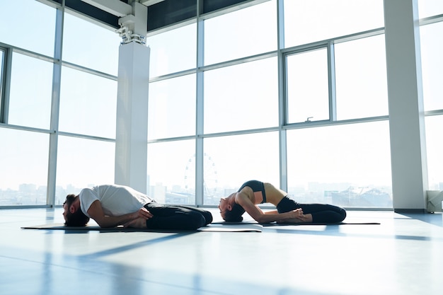 Young sporty man and woman in activewear bending their backs while touching floor with heads during yoga practice