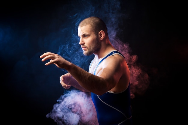 A young sporty man wrestler in a green sports shirt and blue wrestling tights  posing against a blue and red vape smoke background on a black isolated