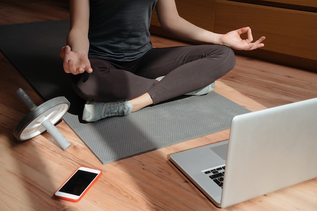 Giovane donna sportiva a praticare yoga online sul pavimento a casa utilizzando laptop e smartphone