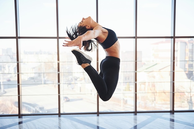 Young sporty woman in sportswear jumping and doing athletic tricks against window in the air.