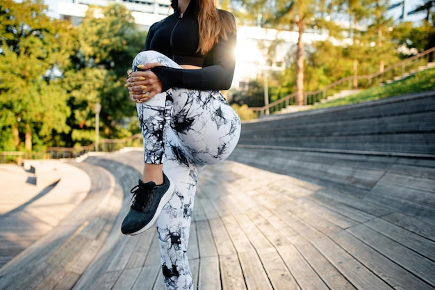 Молодая спортивная женщина в стильной спортивной одежде делает упражнения на растяжку ног на лестнице в парке. Концепция фитнеса и спорта