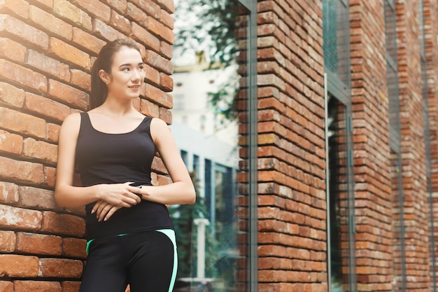 Young sporty woman using smart watch, standing at brick wall background, copy space. Modern technology and fitness concept