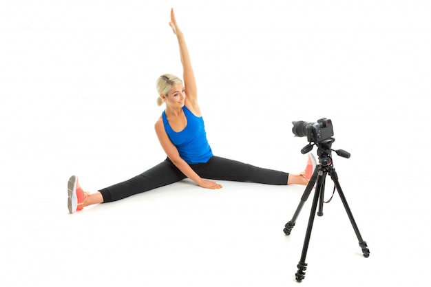 The young sporty woman with a fair hair in a black sports topic, black leggings and bright sneakers does an extension of muscles in front of the camera.