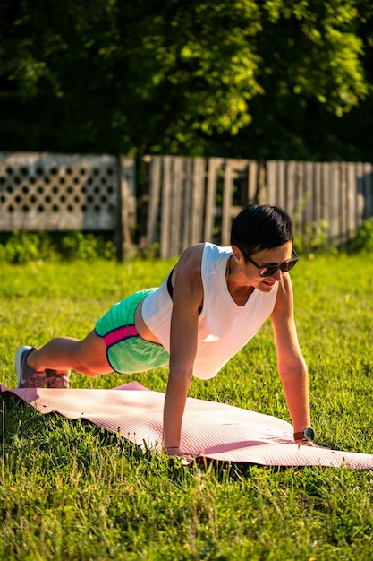 Молодая спортивная женщина с короткими черными волосами делает планку на коврике для йоги на открытом воздухе, тренируется в парке на природе