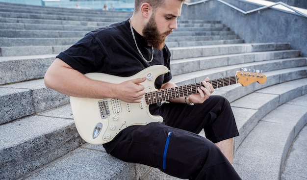 Молодой уличный музыкант играет на гитаре, сидя на гранитных ступенях
