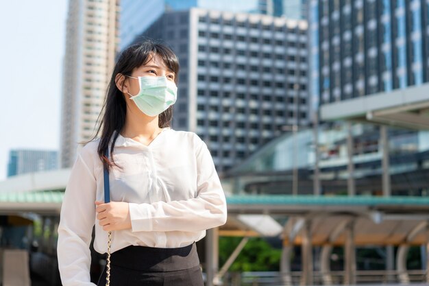 Young stress Asian businesswoman in white shirt going to work in pollution city she wears protection mask prevent dust and COVID-19 with business office building in Bangkok, Thailand.