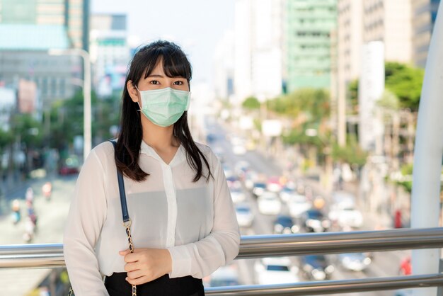 Giovane stress donna d'affari asiatica in camicia bianca che va a lavorare nella città dell'inquinamento, indossa una maschera di protezione per prevenire polvere pm2.5, smog, inquinamento dell'aria e covid-19