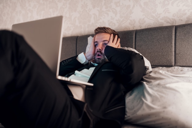 Young stressed businessman lying on the bed in hotel room with laptop in lap and hands on face. Overworking concept.