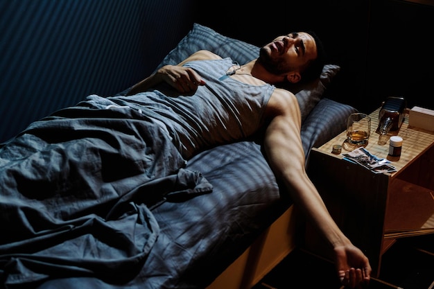 Молодой напряженный мужчина пытается уснуть, лежа на кровати у ночного столика