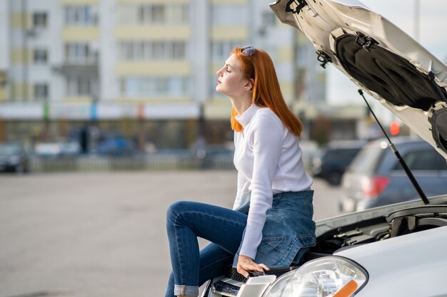 Young stressed woman driver near broken car with popped hood waiting for assistance.