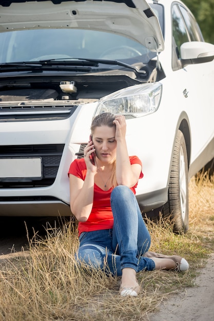 Young stressed woman sitting at the broken car on roadside and talking by phone