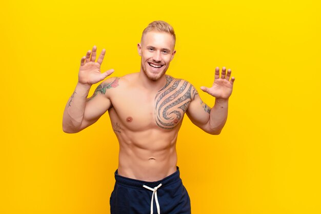 Young strong blonde man screaming in panic or anger, shocked, terrified or furious, with hands next to head against yellow wall