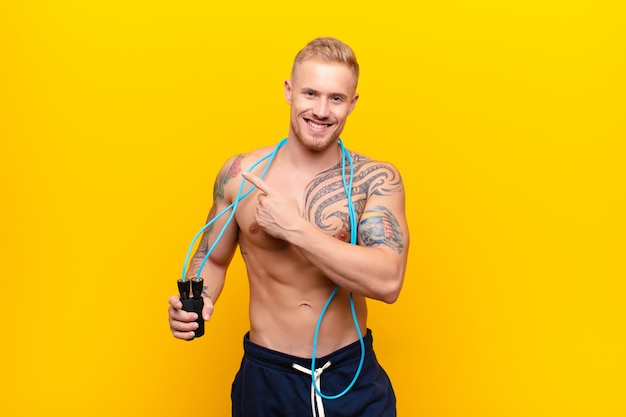 Young strong blonde man smiling cheerfully, feeling happy and pointing to the side and upwards, showing object in copy space with a jump rope