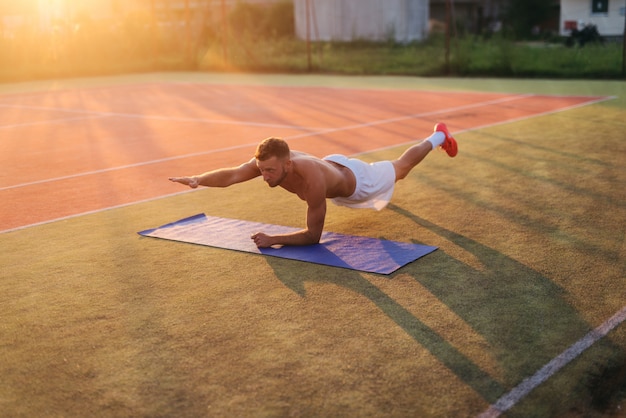 Young strong dedicated man doing crunches on training field. Early summer morning training.