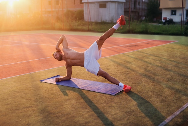 Young strong dedicated man doing crunches on training field. Early summer morning training.