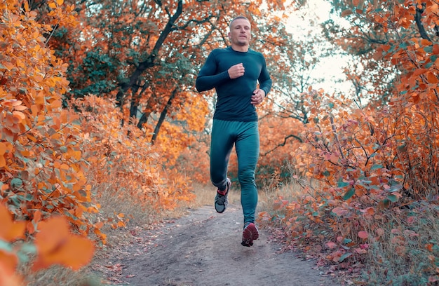 A young strong man in a black sports leggins, shirt and sneakers runs in red autumn forest.