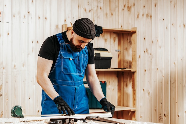 A young strong man builder carpenter works with a wooden bar for making furniture