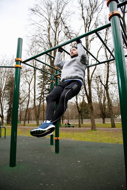 Giovane uomo forte fa pull-up su una barra orizzontale su un campo sportivo in estate in città