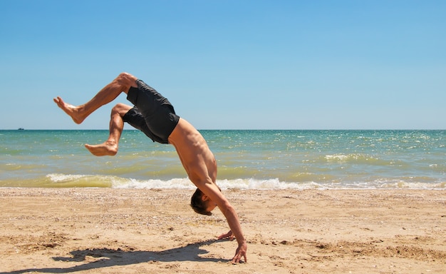 Young strong man in excellent physical shape handstands on the beach near the sea