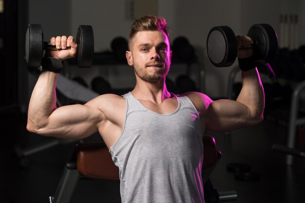 Young Strong Man In The Gym And Exercising Shoulders With Dumbbells  Muscular Athletic Bodybuilder Fitness Model Exercise Shoulder