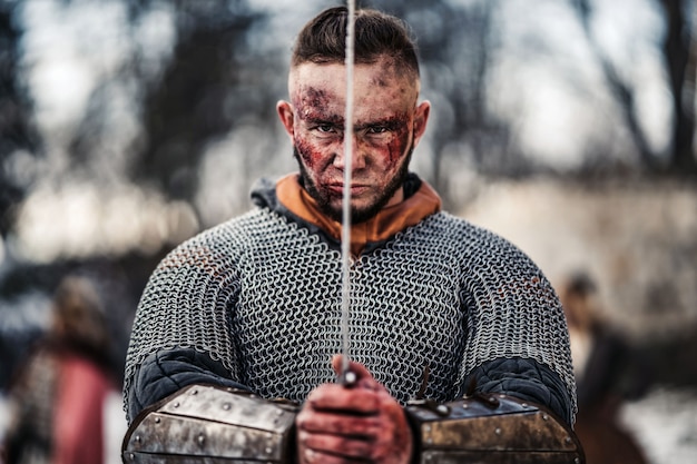 Молодой сильный мужчина в средневековом костюме воина держит меч в руках прямо перед его лицом. Концепция войны и истории