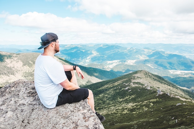 Young strong man sitting on the rock at the top of the mountains peak enjoying the view. hiking concept