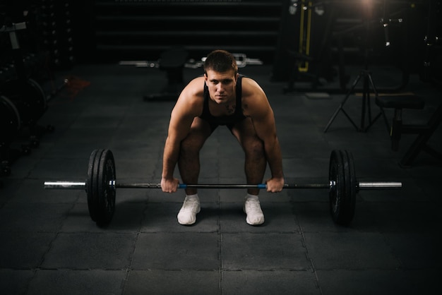 Young strong man with perfect beautiful body wearing sportswear lifting heavy barbell from floor during sport workout training in modern dark gym Concept of healthy lifestyle