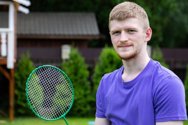 Uomo caucasico di giovane sport forte che posa con la racchetta di badminton sulla natura verde. concetto di gioco amatoriale di badminton, attività estive all'aperto.