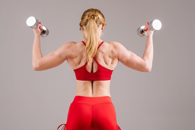 Young strong and sporty woman in red cloth doing exercises with dumbbells on gray background