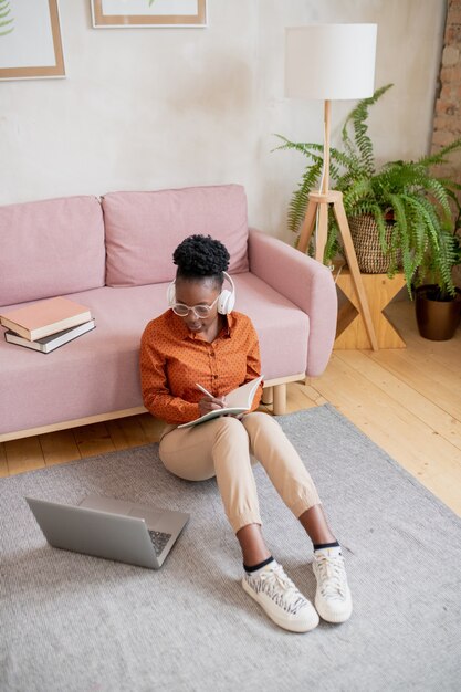 Young student of african ethnicity making notes while sitting by couch