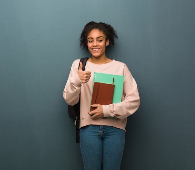 Giovane donna di colore dello studente che sorride e che alza pollice in su. lei tiene libri.