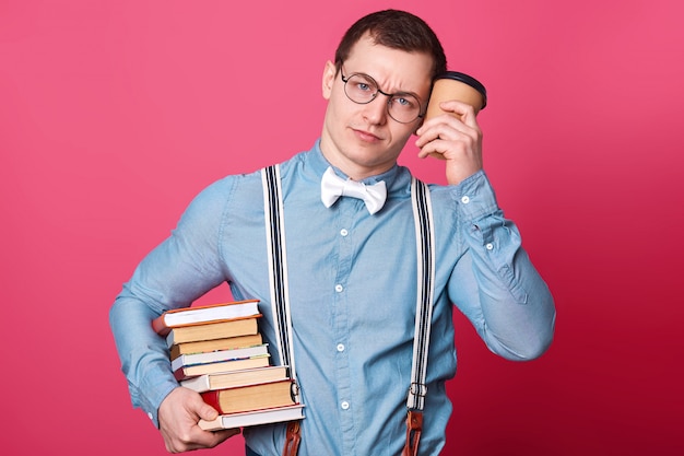 Young student in blue shirt in one tone, looks sleepy and exuasted, holds coffee near head and huge stack of books