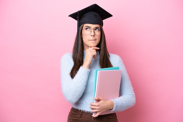 卒業した帽子を身に着けている若い学生のブラジル人女性は、ピンクの背景に疑いと思考を持っています