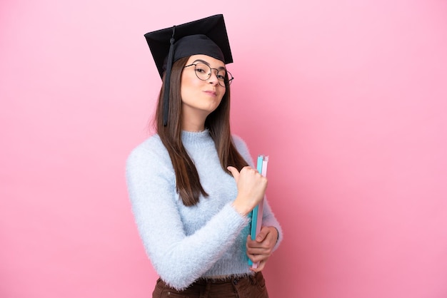 ピンクの背景に分離された卒業帽子を身に着けている若い学生ブラジル人女性は誇りと自己満足