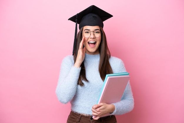 ピンクの背景に卒業した帽子をかぶった若い学生のブラジル人女性が口を大きく開けて叫ぶ