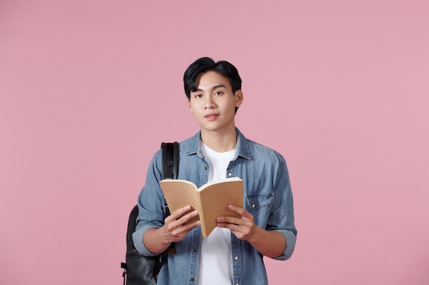 A young student carrying a backpack and holding an open book reading isolated on a pink background