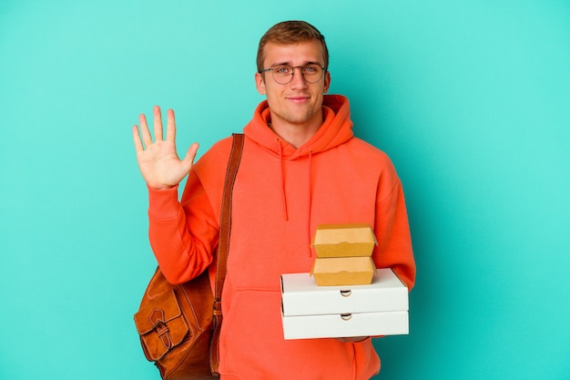 Giovane studente caucasico che tiene hamburger e pizze isolati su sfondo blu sorridente allegro che mostra il numero cinque con le dita.