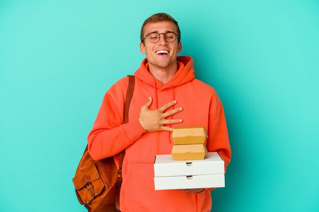 Young student caucasian man holding hamburgers and pizzas isolated on blue laughs out loudly keeping hand on chest.