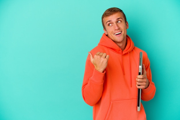 Young student caucasian man holding a laptop isolated on blue background points with thumb finger away, laughing and carefree.