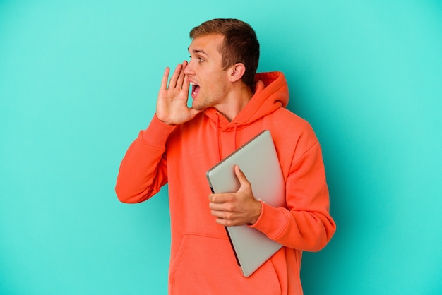 Uomo caucasico del giovane studente che tiene un computer portatile isolato su gridando blu e tenendo il palmo vicino alla bocca aperta.