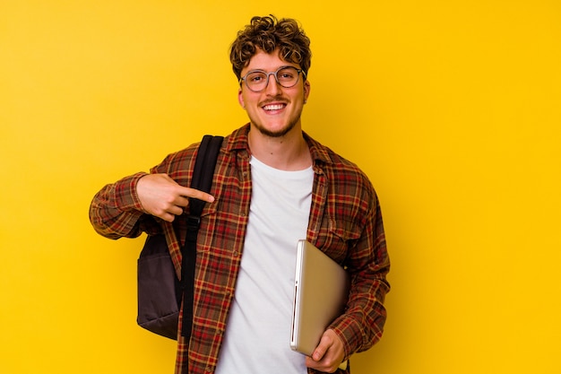 Young student caucasian man holding a laptop isolated on yellow background person pointing by hand to a shirt copy space, proud and confident