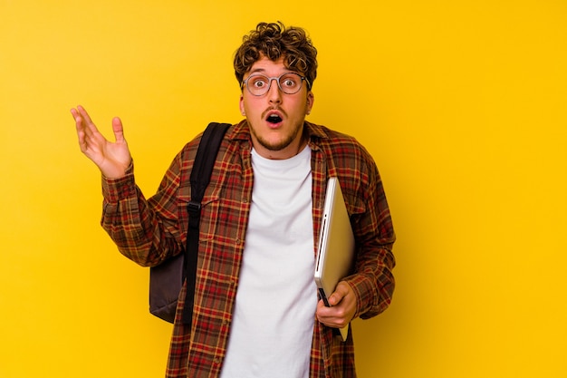 Young student caucasian man holding a laptop isolated on yellow background surprised and shocked.