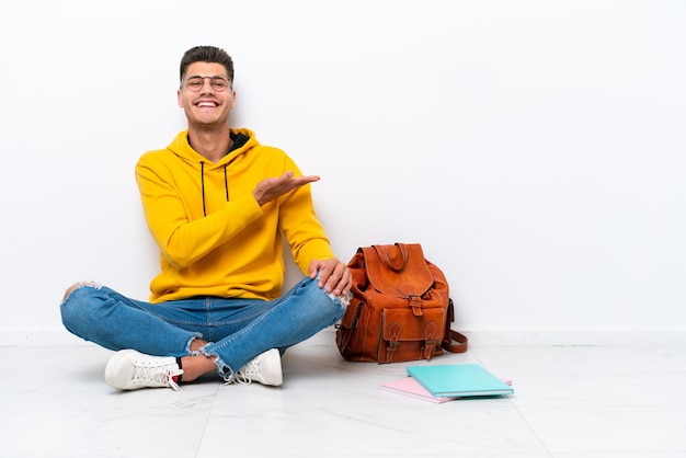 Young student caucasian man sitting one the floor isolated on\
white background presenting an idea while looking smiling\
towards